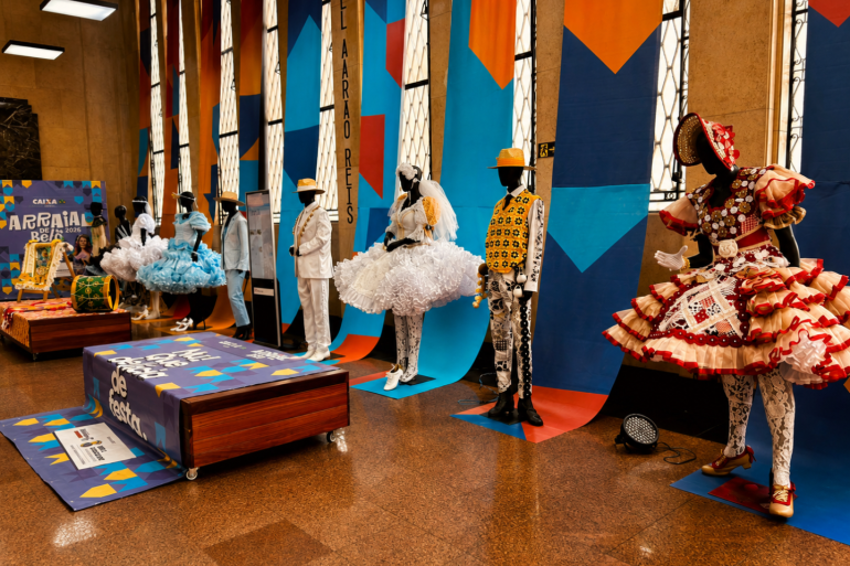 Exposição de figurinos do Arraial de Belô 2026 no hall da Sede da Belotur. Manequins exibem trajes típicos de quadrilhas juninas, com vestidos coloridos, chapéus e roupas ornamentadas. 