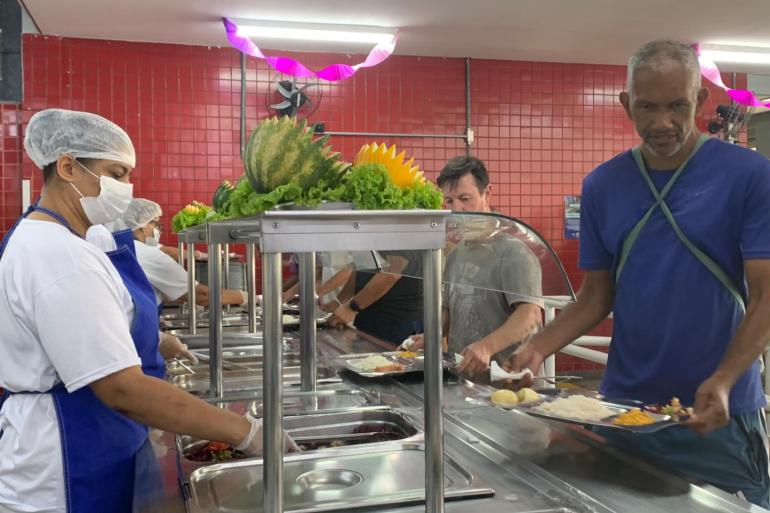 Trabalhadores se servem durante refeição em restaurante popular, com atendimento realizado por profissionais uniformizados no balcão de alimentação.