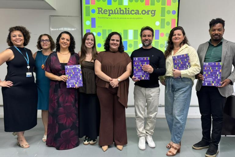 PBH alcança 86% em gestão do desenvolvimento de pessoas, aponta diagnóstico da República.org Um grupo em uma sala de reunião, com oito pessoas adultas em pé, lado a lado, diante de uma tela de apresentação. As pessoas sorriem para a câmera; algumas seguram exemplares de um relatório com capa roxa e colorida. Ao fundo, a tela exibe a identidade visual da República.org, com o texto “Transformar a gestão de pessoas no serviço público” e o ano “2026”, sobre um fundo verde-limão com formas geométricas coloridas.