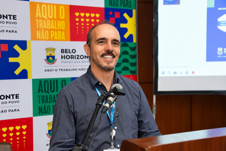 Trilhas de Aprendizagem em Compras Públicas discutem experiências de inovação A foto mostra um homem adulto sorrindo, em pé e falando em frente a dois microfones. Ele usa uma camisa azul-marinho, crachá com cordão azul e óculos pendurados na camisa. Ao fundo, há um painel colorido com o brasão e o nome da Prefeitura de Belo Horizonte, além da frase “Aqui o trabalho não para”. À direita, aparece uma tela de apresentação e, na frente, um púlpito de madeira.