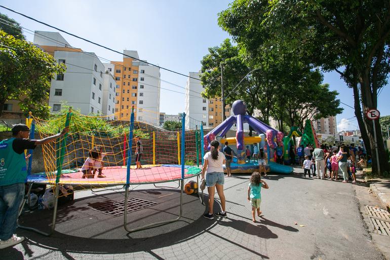 A Rua é Nossa terá jazz, blues e exposição de carros antigos neste domingo (2) Em uma rua fechada ao trânsito, crianças e famílias participam de atividades recreativas ao ar livre. À esquerda, duas crianças brincam em uma cama elástica cercada por uma rede colorida, acompanhadas por um monitor. Ao centro e ao fundo, há grandes brinquedos infláveis em formatos de polvo, dinossauro e melancia, com várias pessoas reunidas ao redor. Uma mulher caminha pela rua de mãos dadas com uma criança. A cena acontece em um dia ensolarado, cercada por árvores e prédios residenciais.