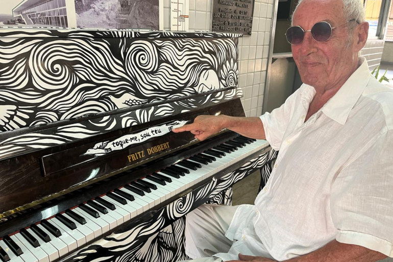 Alberto Lombardi celebra encontro entre Itália e Brasil em espetáculo no Teatro Raul Belém Machado Fotografia do artista Alberto Lombardi, um senhor de pele clara, calça e camisa brancas e óculos escuros redondos. Ele está sentado ao lado de um piano vertical totalmente ilustrado em preto e branco. Alberto aponta para a tampa do instrumento, onde está escrito "toque-me, sou teu" junto à marca Fritz Dobbert. Ao fundo, uma parede clara com placas e fotos decorativas.