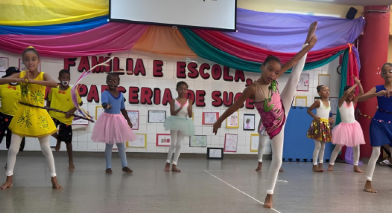 #paratodosverem: crianças se apresentam em um palco escolar decorado com tecido colorido nas cores amarelo, azul, rosa e verde. No fundo, está a frase “Família e Escola, Parceria de Sucesso” e um homem em pé, à esquerda. As crianças usam roupas como vestidos, collants, saias de balé e bermudas. Elas realizam movimentos de dança e ginástica. À direita, pessoas adultas assistem sentadas e aplaudem. 