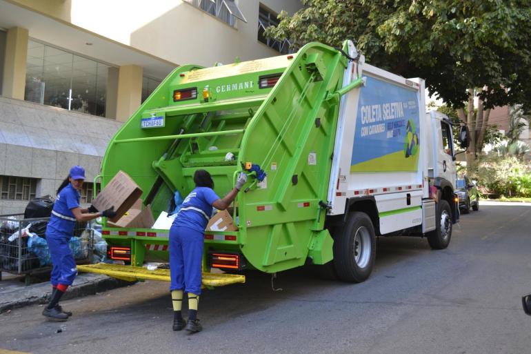 Duas trabalhadoras da limpeza urbana realizam a coleta seletiva porta a porta, colocando materiais recicláveis em caminhão identificado para o serviço.