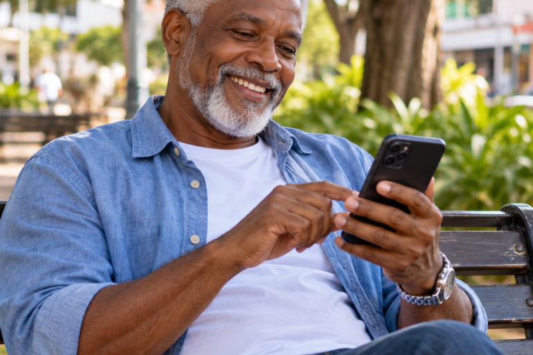 Um homem negro, de aparência idosa, mas com semblante jovem e alegre, está sentado em um banco de praça. Ele tem cabelos curtos grisalhos e barba branca bem aparada. Usa uma camisa azul-clara aberta sobre uma camiseta branca, calça jeans escura e relógio prateado. Ele segura um celular preto com uma das mãos e toca na tela com o dedo da outra, aparentemente lendo ou mexendo no aparelho. Está sorrindo.