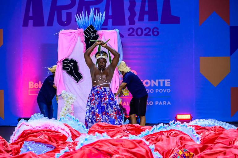 A imagem mostra um homem negro, vestido com uma saia colorida, azul e vermelha, se apresentando em um palco enfeitado com uma manta vermelha