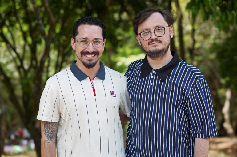 Dois homens brancos posam lado a lado em uma área arborizada, sorrindo para a foto. Gu da Cei veste camisa pólo branca com listras finas e gola azul-marinho; Benedito Ferreira está de camisa azul-marinho com listras verticais brancas e azuis. Ambos usam óculos.