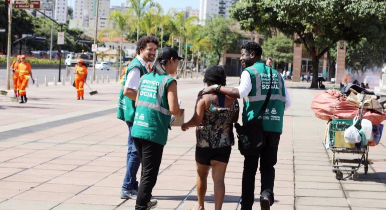 Serviço Especializado de Abordagem Social Foto de três pessoas usando coletes verdes abraçando uma mulher em situação de rua
