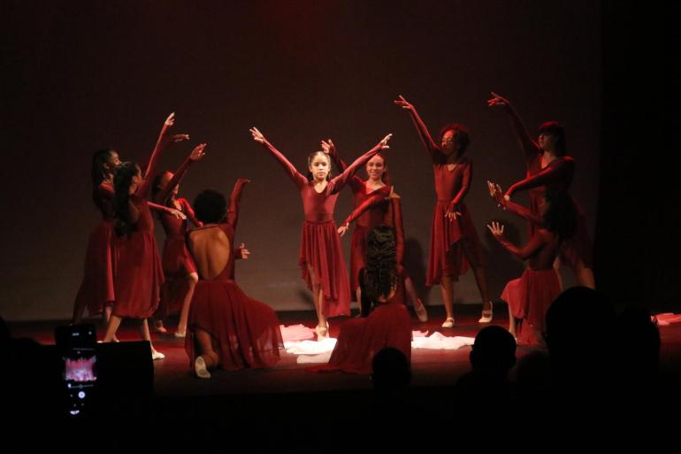 Ballet Val Adriani apresenta trajetória de formação no Terça da Dança Grupo de dançarinas no palco, vestindo vestidos vermelhos, em uma apresentação. Uma bailarina central está com os braços erguidos em forma de V, rodeada por outras que espelham sua pose. A iluminação é dramática, com foco nas bailarinas e o restante do palco escuro.