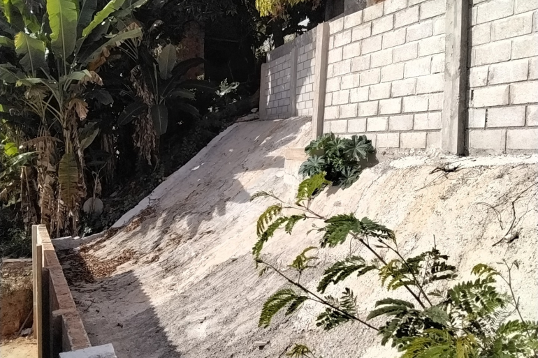 Prefeitura de BH entra na fase final das obras de contenção de risco geológico no Taquaril Estrutura de cimento cinza claro feita para contenção de encosta com risco de desabamento, com plantas de médio e grande porte ao fundo.