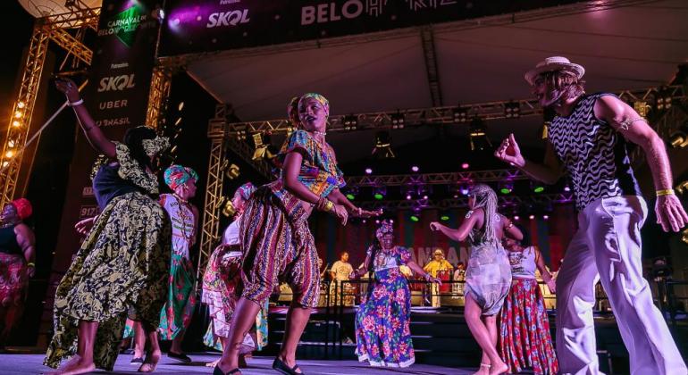 Grupo musical com sete pessoas com vestimentas de inspiração afro se apresentam no palco do Carnaval de Belo Horizonte. 