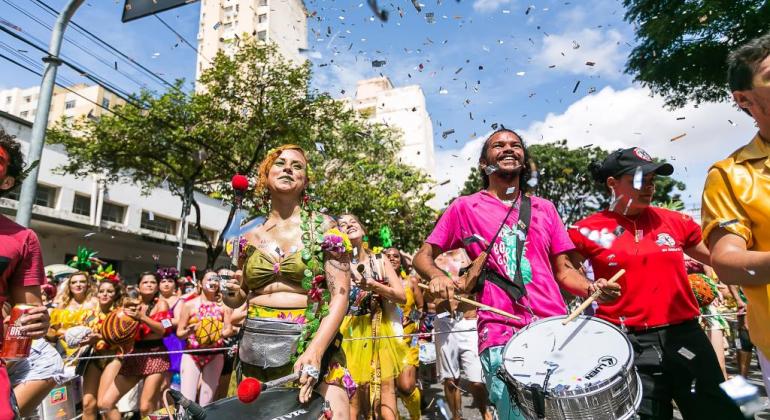 Integrantes de bloco de rua tocam instrumentos de tambor em dia de sol