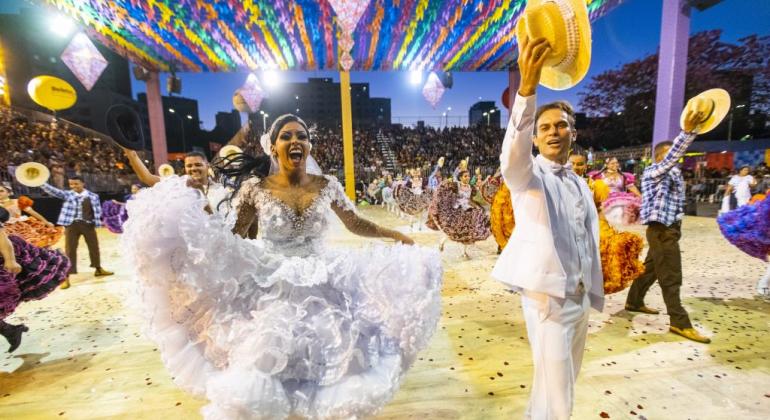 Foto: Bruno Figueiredo Noiva e noivo caipiras sorriem e acenam para a plateia, com outros casais da quadrilha ao fundo, em apresentação, à noite.