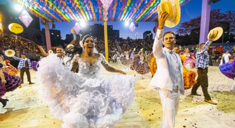 Noiva e noivo caipiras sorriem e acenam para a plateia, com outros casais da quadrilha ao fundo, em apresentação, à noite. 