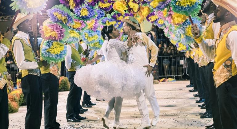 Noivo e noiva de quadrilha junina se beijam, cercados por homens e arranjo de flores. 