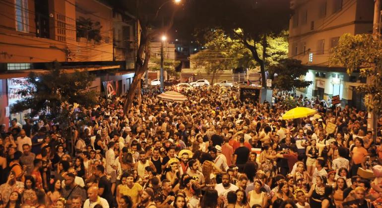 Rua com mais de quinhentas pessoas pulando carnaval, durante a noite. 