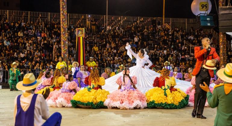 Casais se apresentam em uma dança de quadrilha, com público ao fundo.