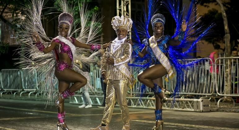 Rei Momo e Rainha e Princesa do Carnaval sambam em avenida, à noite. 