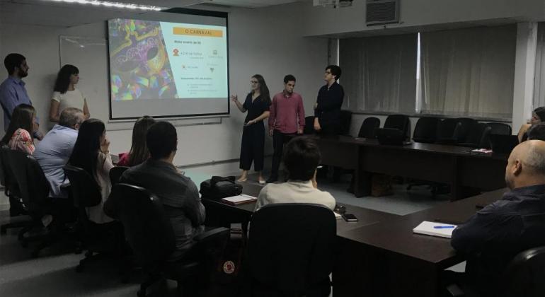 Foto: Divulgação PBH Nove adultos sentados em sala com mesa em forma de "u", e três em pé, fazendo exposição, ao lado de projeção em imagem.