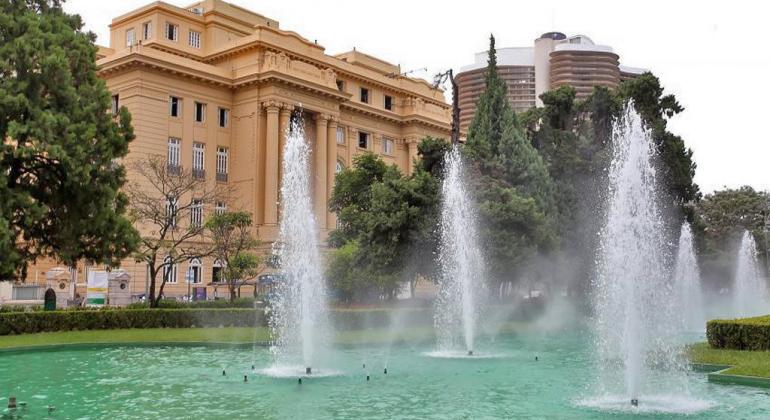 Fonte de água da Praça da Liberdade com prédio histórico ao fundo, durante o dia. 