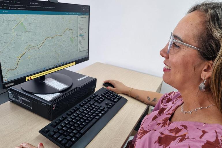 Uma mulher está sentada diante de um computador, em um ambiente de escritório claro e organizado. Ela aparece de perfil, sorrindo enquanto olha para a tela e segura um mouse com a mão direita. Na tela, o site do BHmap.