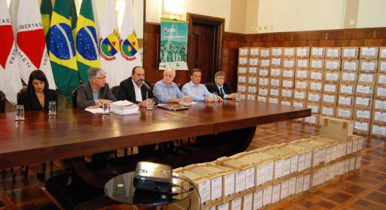 O prefeito de Belo Horizonte, Alexandre Kalil, junto a outros cinco membros da equipe municipal, sentados em uma mesa no Salão Nobre da PBH. à frente, mais de cem caixas com arquivos da BHTrans. 