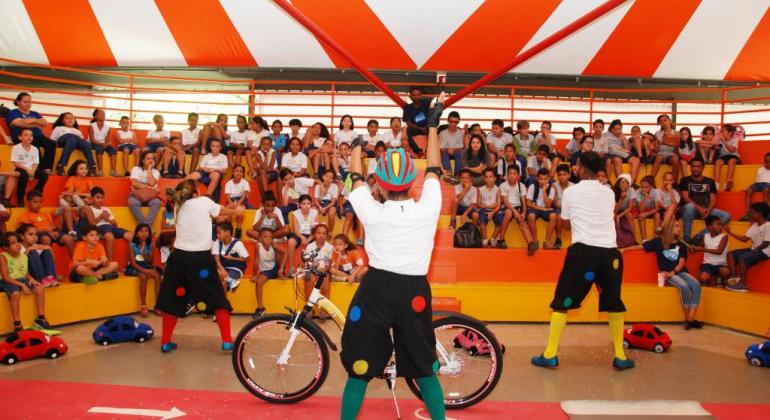 Circo Transitando Legal leva diversão e aprendizado a estudantes municipais Foto de três artistas em cena e cerca de cinquenta crianças sentadas em plateia