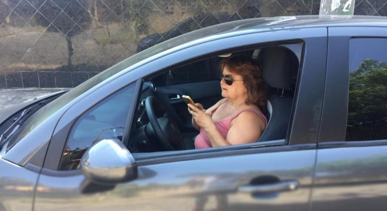 Motorista feminina, com carro estacionado e janela aberta, confere aplicativo no celular, durante o dia.