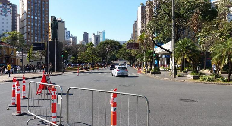 Avenida com cones e grades de interdição e carros passsando ao fundo, durante o dia. 