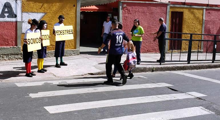 Três pessoas, vestidas de palhaço, na calçada, em frente a um local de travessia de pedestres, com três placas que formam o dizer: "somos todos pedestres". Na travessia, um adulto atravessa com uma criança. 