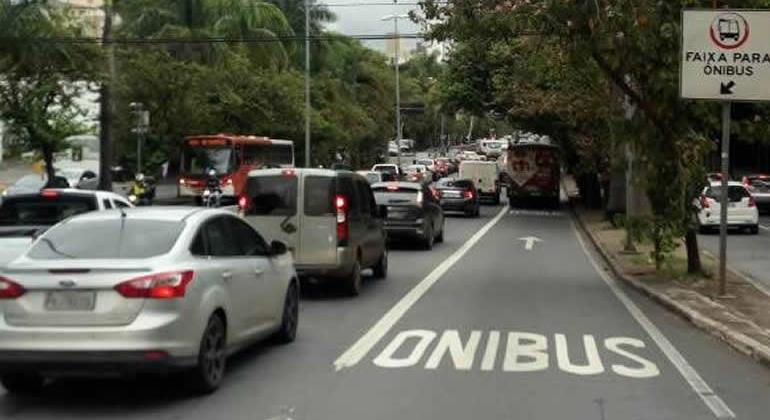 Avenida com carros à esquerda e pista exclusiva de ônibus à direita, com ônibus ao fundo. 