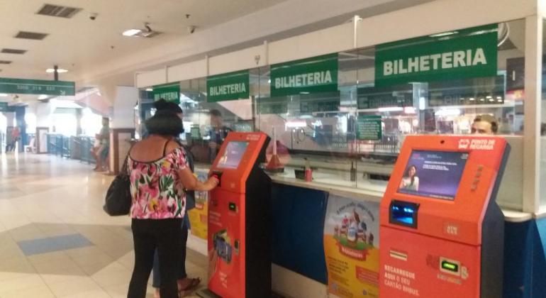 Duas mulheres utilizando máquina de ponto de recarga do cartão BHBus, próximo a bilheterias.
