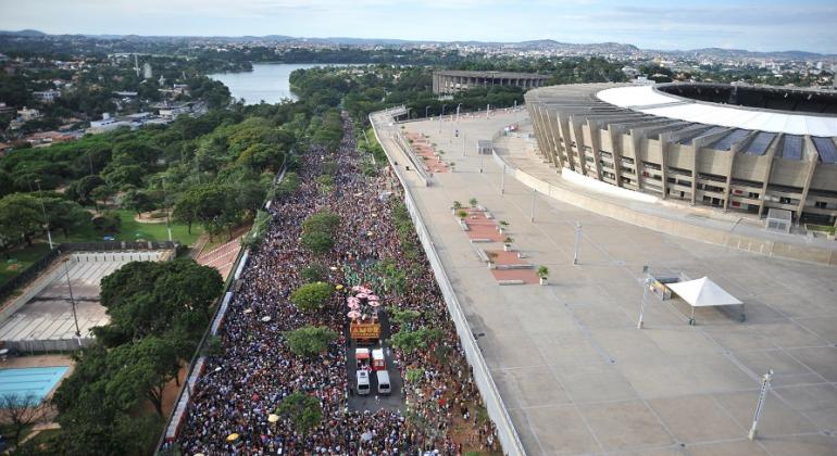 Bloco de rua com mais de quinhentas pessoas em avenida, ao lado do Mineirão, tendo ao fundo o Mineirinho e a Lagoa da Pampulha, durante o dia. 