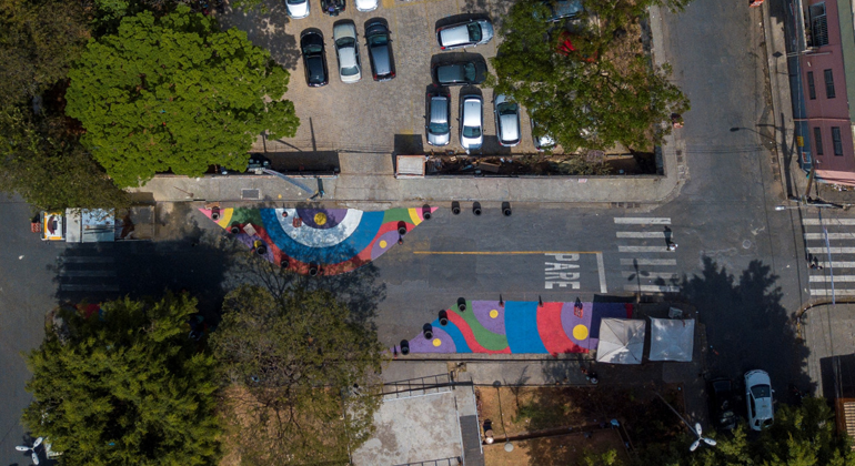 Imagem aérea de uma rua com pinturas e um estacionamento de carros ao lado, durante o dia. 