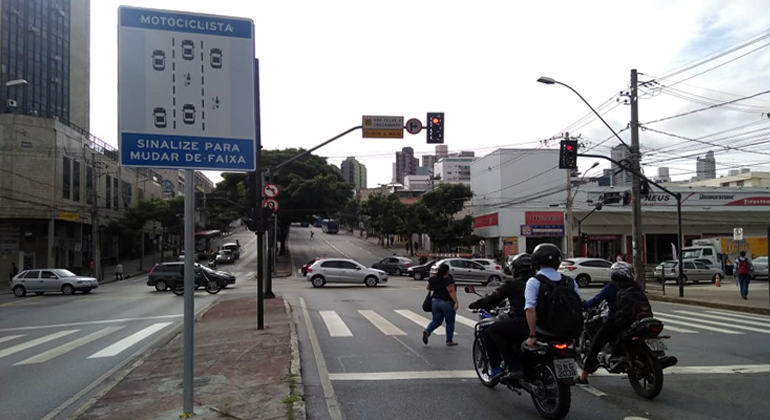 Motociclistas em avenida, ao lado de placa, onde está escrito: "Motociclista: sinalize para mudar de faixa". 