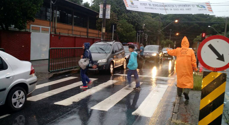 Agente de trânsito com capa de chuva acompanha alunos que atravessam a faixa de pedestre em direção à escola, durante o dia. 