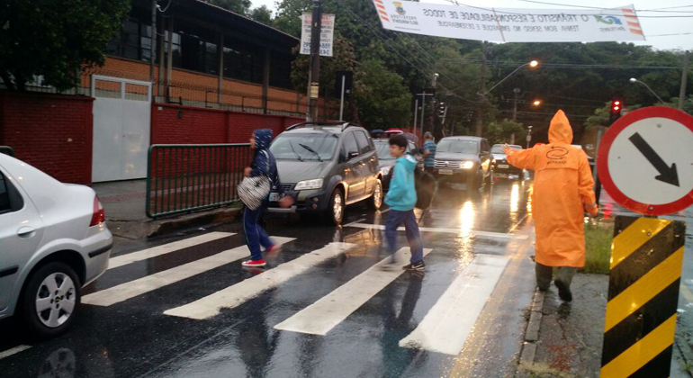 Foto: Divulgação PBH Agente de trânsito com capa de chuva acompanha alunos que atravessam a faixa de pedestre em direção à escola, durante o dia.