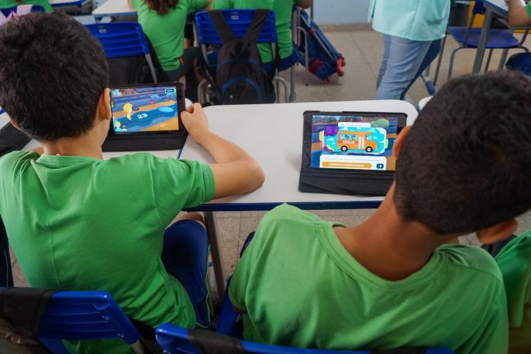 BHTrans promove ações educativas de mobilidade para crianças e adolescentes no Festival do Programa Saúde na Escola Dois estudantes de uniforme verde e azul, sentados de costas para a câmera, usam tablets em uma sala de aula de uma escola no Brasil. O tablet do aluno da esquerda exibe o jogo de aventura de pular em poças 'Pega-poça', e o do aluno da direita mostra o jogo da escola. Os alunos estão cercados por outros estudantes e cadeiras e mesas brancas e azuis.