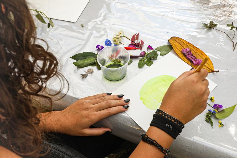 Museus Pampulha destaca o fazer artesanal, a memória e a paisagem urbana na programação de agosto Com foco nas mãos de uma mulher a foto mostra uma mesa branca com flores e folhas em cima, um pote de tinta e uma pintura sendo feita pela mulher.