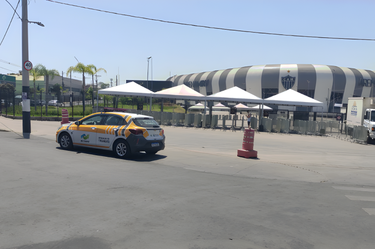 Operação de trânsito para Atlético x Grêmio na Arena MRV, neste domingo (16) A imagem mostra uma área externa pavimentada em dia ensolarado, com destaque para a Arena MRV ao fundo, o estádio do Clube Atlético Mineiro. No primeiro plano e ao centro, há movimentação de apoio e trânsito.