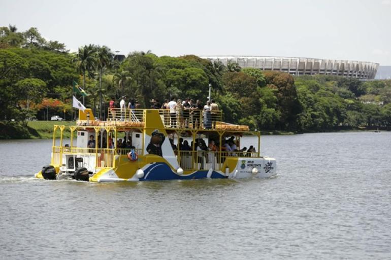 Foto de um barco com tonalidade amarela, azul e branca tansportando pessoas em navegação por espelho d'água, com o Mineirão ao fundo