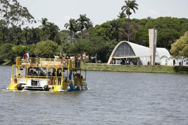 A imagem mostra o Capivarã, embarcação turística de cores branca e amarela, navegando pela Lagoa da Pampulha, em Belo Horizonte. Há passageiros a bordo, distribuídos nos dois pavimentos do barco. Ao fundo, aparece a Igreja São Francisco de Assis, um dos principais marcos arquitetônicos do Conjunto Moderno da Pampulha, cercada por árvores e palmeiras. O dia está claro, com céu parcialmente nublado, e a paisagem destaca a integração entre a lagoa, a vegetação e a arquitetura modernista.