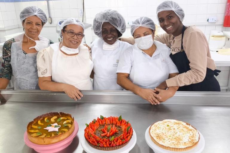 Cinco mulheres de avental e touca em uma cozinha com três tortas doces na mesa. 