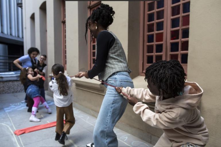 Programação do “Museus Centro” celebra o Mês das Crianças com atividades para todas as idades Crianças e adultos participam de uma brincadeira com uma corda vermelha em um corredor externo. Em primeiro plano, à direita, uma criança de cabelos crespos e conjunto bege segura a corda com as duas mãos e inclina o corpo para trás. Ao centro, uma adulta de tranças presas, colete xadrez e calça jeans também segura a corda. Outras participantes aparecem ao fundo. Há desenhos de giz no piso e janelas rosadas com vidros coloridos na parede.