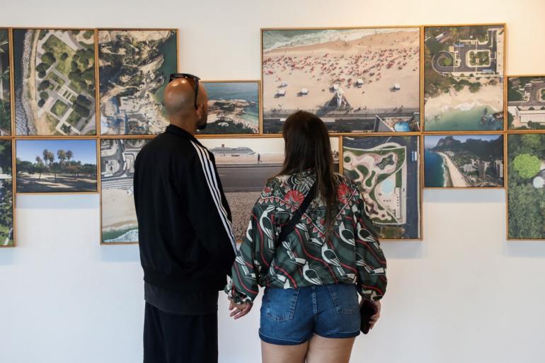 Na foto, um casal observa uma parede de quadros com fotografias da exposição "Trans:paisagem". Um homem e uma mulher estão de costas e à sua frente está uma parede branca com uma série de quadros com fotos dos jardins de Burle Marx.