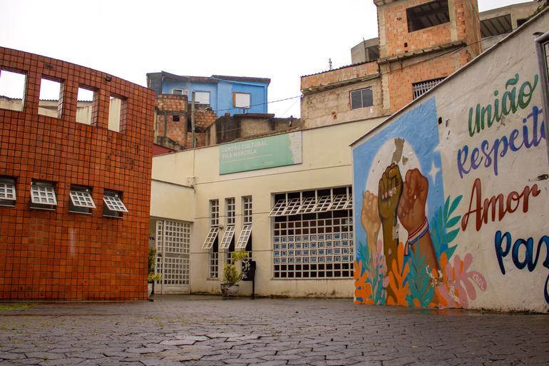 Mês de agosto terá atrações variadas nos centros culturais São Geraldo, Alto Vera Cruz e na Usina de Cultura Foto do Centro Cultural Vila Marçola. A imagem mostra o pátio de entrada, a fachada do equipamento cultural, casas ao fundo e, à direita, um mural colorido com mãos erguidas e a frase "União, respeito, amor e paz".
