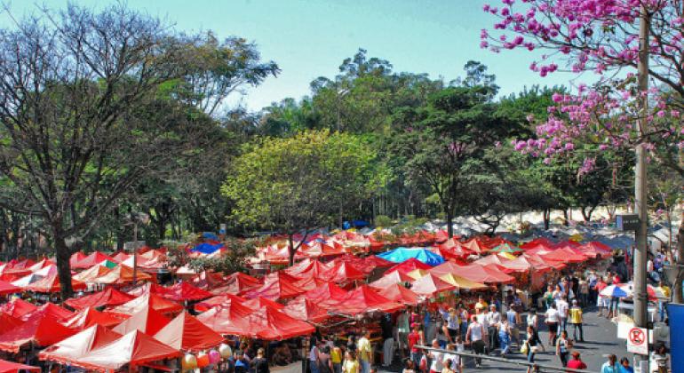 Mais de oitenta barraquinhas vermelhas na Feira de Artesanato da Afonso Pena, com pessoas passeando, durante o dia. 