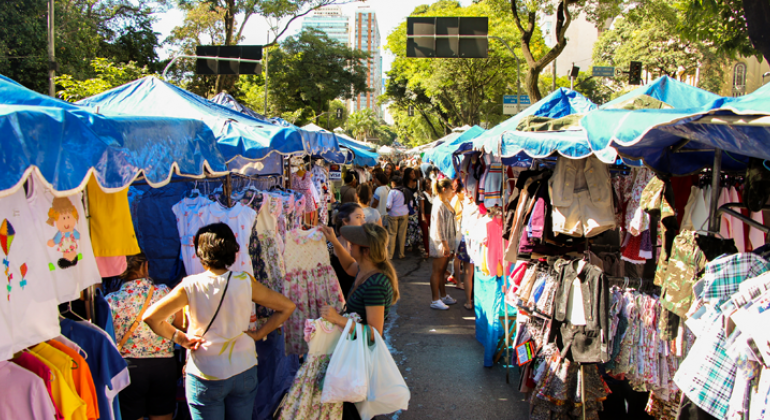 Duas filas de barracas de artesanato da feira de Avenida Afonso Pena, com roupas nas barracs e pessoas transitando entre as barracas, durante o dia. 