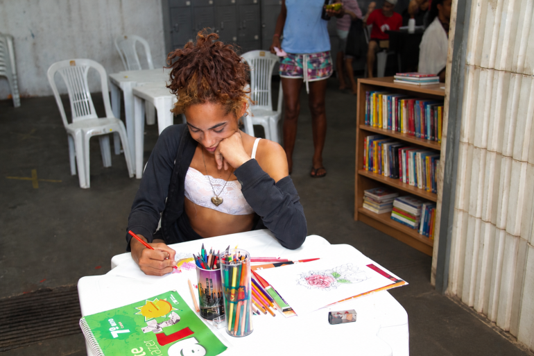 Centro Pop Mulheres promove campanha de arrecadação de livros para ampliar acesso à leitura de atendidas pelo serviço Uma pessoa de cabelos cacheados castanhos, presos no alto da cabeça, está sentada a uma mesa branca, desenhando com um lápis. Sobre a mesa há desenhos, lápis de várias cores, materiais escolares e um caderno verde. Ao fundo, aparecem mesas e cadeiras brancas, outras pessoas e uma pequena estante de madeira com livros coloridos.