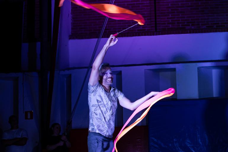  Em um palco sob iluminação azul, um artista circense sorri enquanto movimenta duas fitas coloridas que se desenrolam no ar. Ele segura cada fita por uma haste